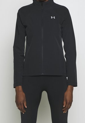 Personne portant une veste zippée noire ajustée et des leggings noirs texturés, mains relâchées le long du corps sur un fond uni.