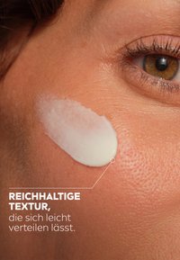 Cremige Lotion auf der Haut, glatte Textur, weiße Farbe, leicht verteilbar, wird auf gebräunter Haut mit sichtbaren Poren und feinen Härchen aufgetragen.