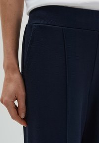 Marinebroeken van een textuurstof, met een hoge tailleband, zijzakken en voorplooien. Hand die op de zijkant van de stof rust.