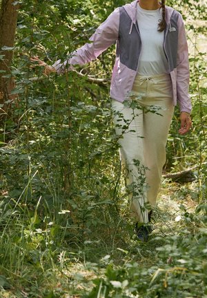 Osoba idąca przez gęsty zielony las, ubrana w jasne spodnie, białą koszulę i fioletową kurtkę outdoorową, dotykająca roślin wzdłuż ścieżki.