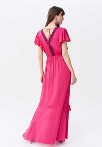 Langes rosa Kleid mit schwingenden Ärmeln, V-Ausschnitt hinten und elastischem Bund. Besitzt eine Spitzenverzierung entlang des Rückens und eine fließende Silhouette.