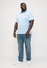 Camisola polo azul claro com um padrão texturizado, mangas curtas e colarinho, combinada com jeans azul claro e ténis brancos.