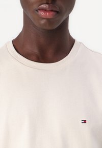 Nærbilde av en person som har på seg en ensfarget beige genser med rund hals og en liten rød, hvit og blå rektangulær logo på brystet.