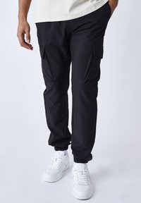 Pantalones cargo negros con puños elásticos, que cuentan con dos bolsillos laterales y detalles texturizados sutiles. Combinados con zapatillas blancas para generar contraste.