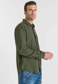 Man in een olijfgroen overhemd met lange mouwen en blauwe jeans, staand zijwaarts tegen een effen lichte achtergrond.