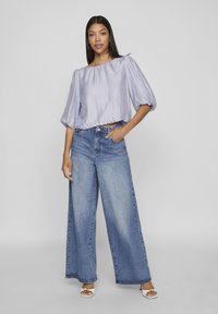 Bluză satinată lila cu umeri la vedere și mâneci bufante, purtată cu blugi din denim albastru, cu talie înaltă și picioare largi. Modelul stă cu mâinile pe șolduri.
