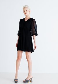 Robe noire en tissu léger, avec des manches bouffantes, une taille cintrée et un col rond agrémenté d'un bouton décoratif.