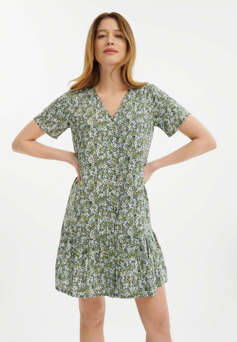 Grünes Blumenkleid mit V-Ausschnitt, kurzen Ärmeln und gerafftem Rock. Es hat ein Muster aus kleinen weißen und blauen Blumen auf einem blätterreichen Hintergrund.