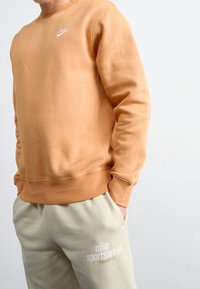 Man med ljusbrun Nike-tröja och beige sweatshirtbyxor med texten "nike sportswear" mot en enkel bakgrund.