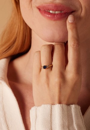 Goldring mit einem ovalen blauen Stein, flankiert von zwei kleineren Steinen. Hand, die am Kinn ruht, vor einem hellen Hintergrund.