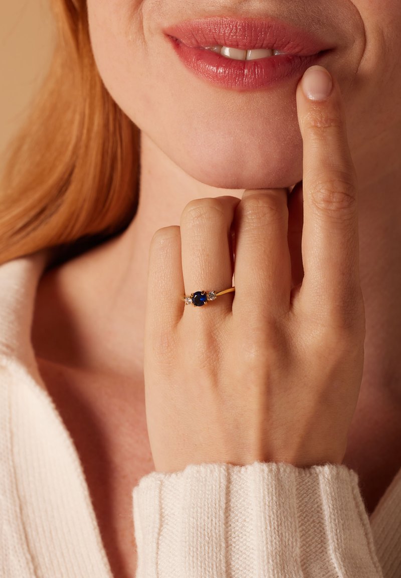 Gouden ring met een ovale blauwe steen, omringd door twee kleinere stenen. Hand rustend op de kin, tegen een lichte achtergrond.