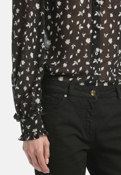 Black floral blouse with white leaf patterns, sheer fabric, and ruffled cuffs, paired with black jeans featuring a gold button detail.