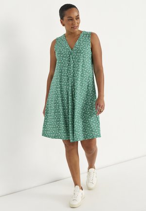 Vrouw loopt, draagt een mouwloze groene bloemenjurk tot kniehoogte en witte sneakers tegen een effen witte achtergrond.