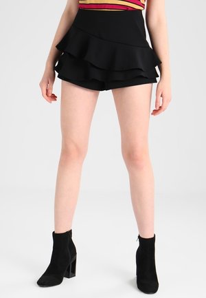 Femme portant un short noir à volants superposés, des bottines noires à talons et un haut rayé sans manches, debout sur un fond blanc.