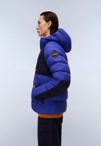 Giubbotto imbottito con un corpo blu brillante e dettagli neri, dotato di cappuccio e di una toppa ricamata sulla manica. Tasche laterali con zip.