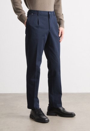 Man draagt marineblauwe op maat gemaakte broek en zwarte leren nette schoenen, staande met handen in de zakken op een lichtgrijze vloer.