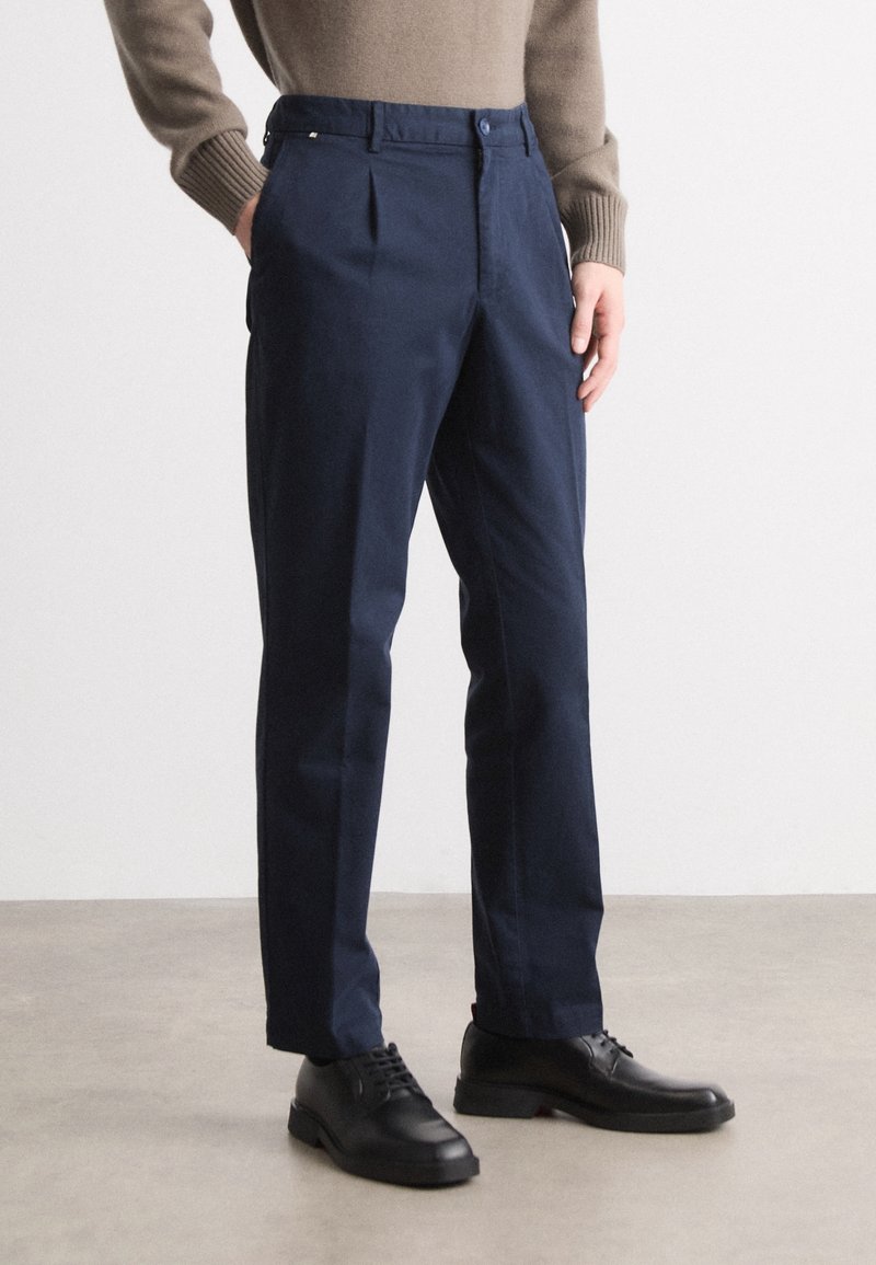 Homme portant un pantalon bleu marine sur mesure et des chaussures en cuir noir, debout les mains dans les poches sur un sol gris clair.