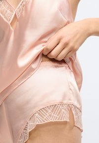 Siidi-sarnane heleroosa pidžaamakomplekt, mis sisaldab V-kaelusega särk ja pitsiga ääristatud lühikesi pükse, millel on geomeetriline muster äärel.