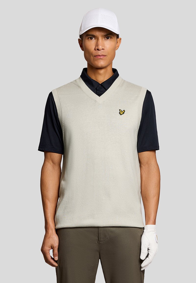 Männlicher Golfer mit weißer Kappe, beigem ärmellosem Pullover über schwarzem Polohemd, brauner Hose und weißem Golfhandschuh an der rechten Hand.