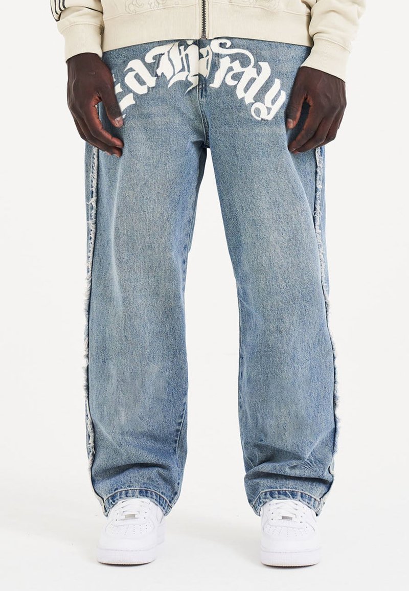 Jeans de mezclilla azul claro con un ajuste relajado, con texto gráfico blanco y paneles laterales desgastados. Combinados con zapatillas blancas.