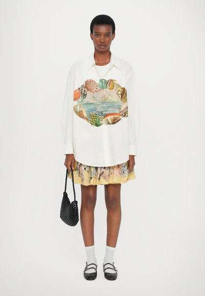White long-sleeve shirt with a colorful shell and landscape graphic, paired with a yellow floral skirt, black handbag, and grey socks.