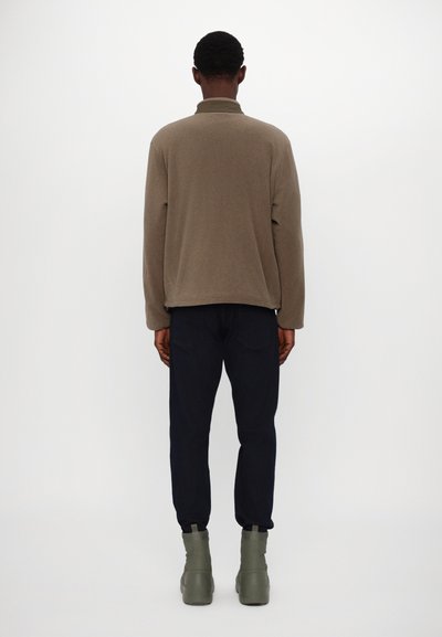 Beige Fleecejacke mit hohem Kragen, langen Ärmeln, kombiniert mit dunkelblauen Hosen und hellgrünen Knöchelboots, von hinten betrachtet.