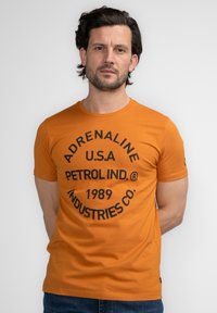 Camiseta de algodón naranja con estampado circular negro que lleva el texto "ADRENALINE U.S.A. PETROL IND.® 1989 INDUSTRIES CO." Manga corta.