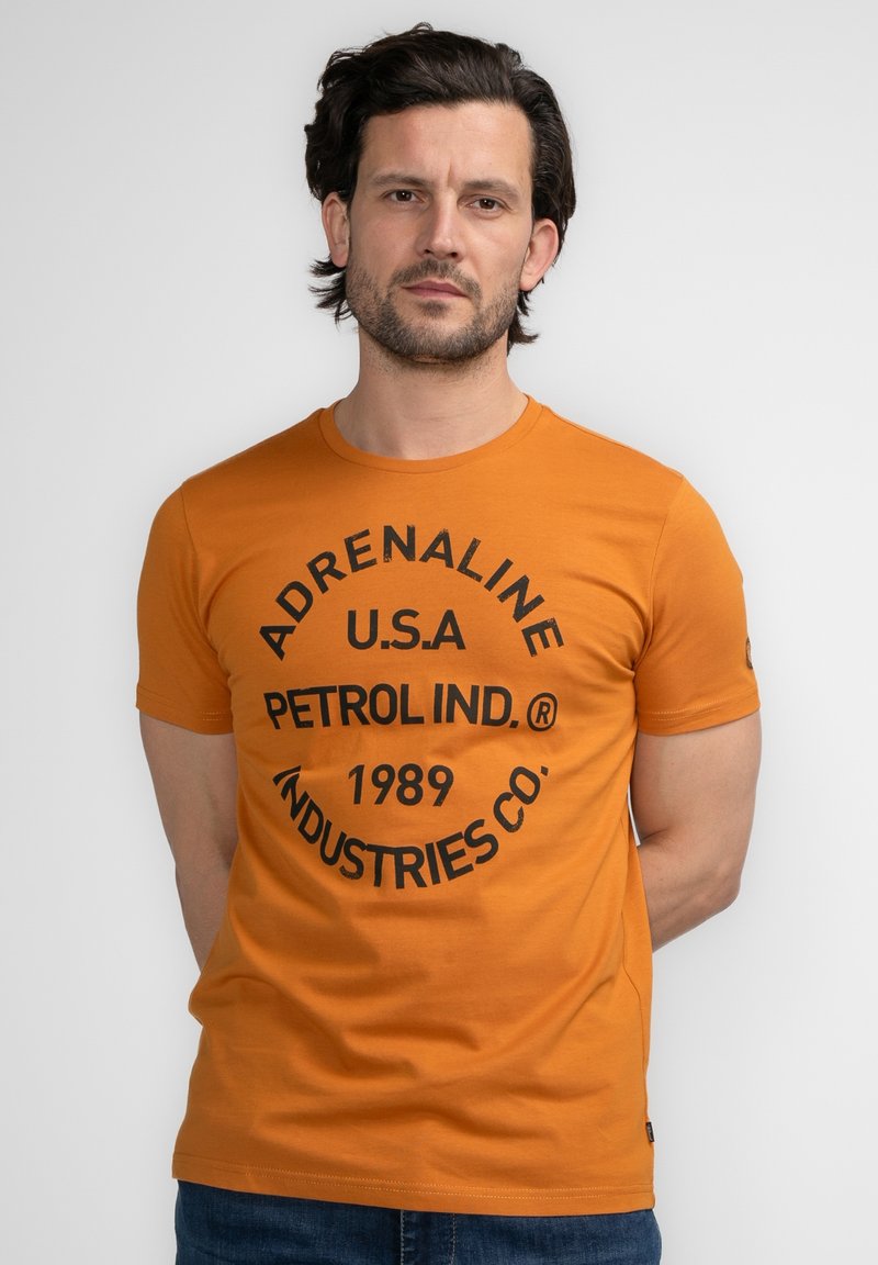 Camiseta de algodón naranja con estampado circular negro que lleva el texto "ADRENALINE U.S.A. PETROL IND.® 1989 INDUSTRIES CO." Manga corta.
