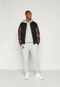 Vīrietis valkā melnu universitātes žaketi ar brūnām ādas piedurknēm, pelēku kapuci un sporta bikses ar zilu "LA" logo, baltas kedas, stāvot uz baltas sienas fona.