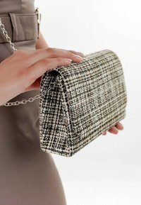 Ręka trzyma torebkę z fakturowanej tkaniny tweed z srebrnym łańcuszkiem w zestawieniu z neutralnym strojem na białym tle.