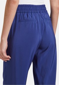 Blå satinbyxor med resårmidja, med två bakfickor och en mjuk textur, visade från sidan.