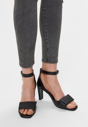 Black heeled sandals with ankle straps, featuring a square toe and textured black leather panels. The heel height is medium.