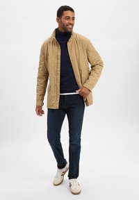 Beige tikattu takki, jossa on pystykaulus, napit ja sivutaskut, päällä tummansininen villapaita ja tummat farkut.