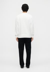 Άνδρας με σγουρά μαύρα μαλλιά, φορώντας απλή λευκή μπλούζα με μακριά μανίκια και μαύρο παντελόνι, στέκεται γυρισμένος προς τα πίσω σε λευκό φόντο.