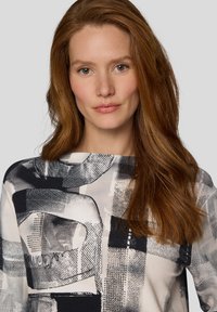 Frau mit langen roten Haaren, die ein schwarz-weiß-grau gemustertes Oberteil mit abstraktem Design trägt, frontal vor einem schlichten Hintergrund steht.