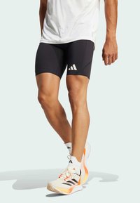 Schwarze Sportshorts mit figurbetontem Design, die drei weiße vertikale Streifen aufweisen. Ein Paar leichte Laufschuhe in Creme und Orange.