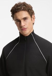 Μαύρο μπουφάν με φερμουάρ και λευκές ραφές. Κατασκευασμένο από υφασμάτινη υφή, διαθέτει ψηλό γιακά και ανάγλυφο λογότυπο στο στήθος.