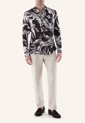 Mann trägt ein schwarz-weiß geblümtes Hemd mit Knöpfen, cremefarbene Hose und dunkelbraune Tasselloafer, steht vor einem schlichten Hintergrund.