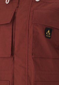 Burgundy jacka tillverkad av slitstarkt tyg; har två framfickor, justerbar dragsnodd och en broderad logotyp på vänster sida.