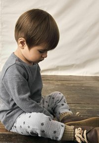 Chemise à manches longues gris côtelé, pantalon gris à motifs, chaussures marron. Assis sur un parquet, la tête penchée, montrant des textures douces et des couleurs sourdes.