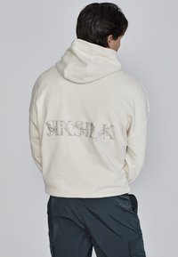 Cremefarbene Kapuzenjacke mit einem detailliert gestickten Logo auf dem Rücken, das kunstvolle florale Elemente aufweist. Weicher, strukturierten Stoff. Lässiger Schnitt.