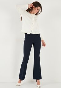 Weißer Strickcardigan mit fransigem Saum, rundem Ausschnitt und Knopfverschlüssen, kombiniert mit navyblauen Schlaghosen und weißen Schuhen mit Kappen.