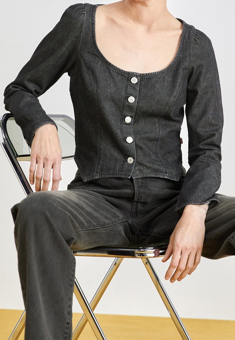 Top en denim noir court à manches longues, décolleté carré et cinq boutons contrastants. Porté avec un pantalon en jean sombre assorti, assis sur une chaise en chrome.