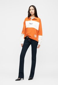 Γυναίκα που στέκεται φορώντας πορτοκαλί και λευκό φούτερ, σκούρο μπλε τζιν, μαύρη ζώνη και μαύρες μυτερές γόβες σε απλό λευκό φόντο.