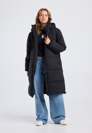 Manteau noir en duvet avec une capuche, présentant un design matelassé et une fermeture éclair à l'avant. Associé à un jean large bleu clair et des baskets blanches.