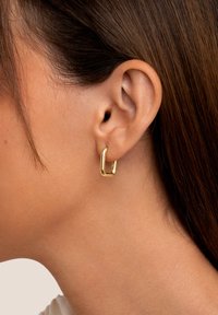 Nahaufnahme eines Ohrs einer Frau mit glattem braunem Haar, die einen kleinen, dicken, rechteckigen goldenen Kreolenohrring trägt.