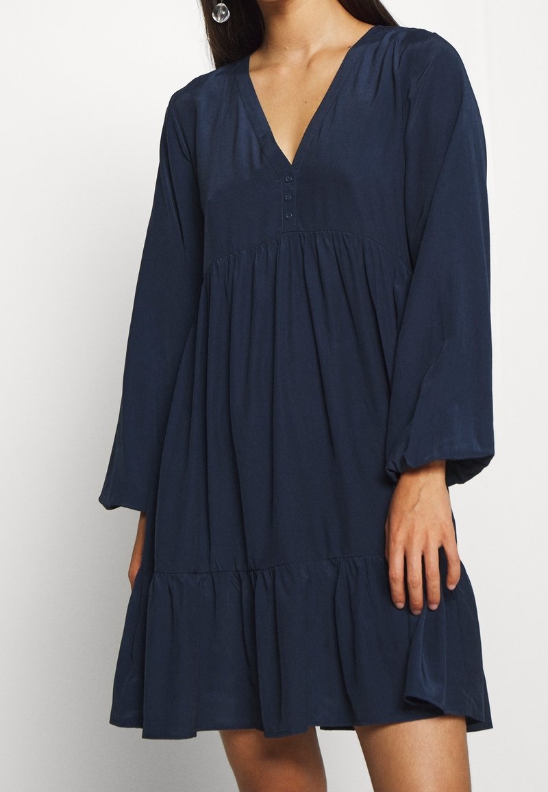Mörkblå klänning med V-ringning, långa ärmar och en lager på lager-ruffelavslutning. Tillverkad av mjukt tyg med knappdetaljer på bysten.