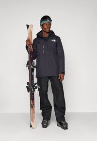 Muž v čiernej lyžiarskej bunde a nohaviciach, drží pár lyží, má na nohách lyžiarky, na čele lyžiarske okuliare a zelenú čiapku.