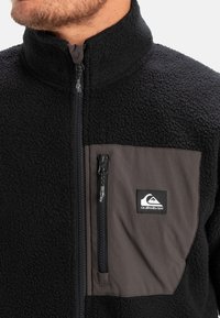 Mustan fleece-jakkiga kõrge kaelusega, ees asuva tõmblukuga ja halli rinna taskuga. Omab logopulka ja kontrastseid tekstuure.