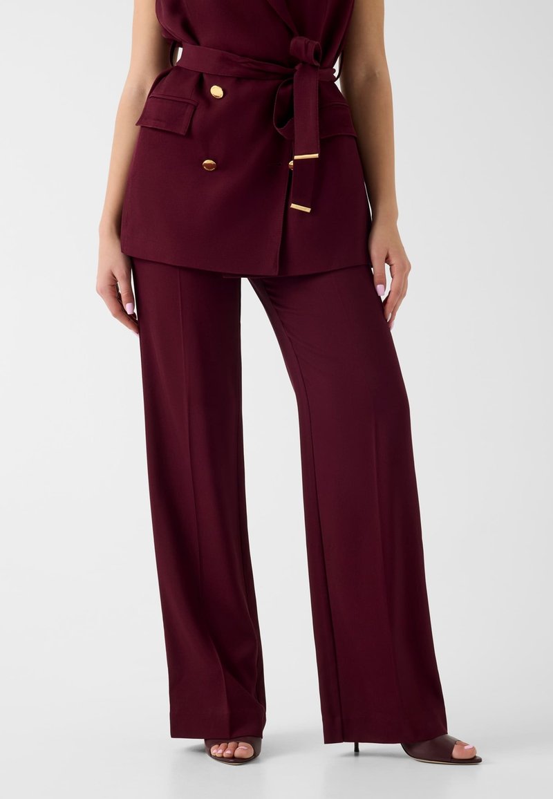 Femme portant un pantalon bordeaux à jambes larges avec un blazer ceinturé assorti orné de boutons dorés, se tenant debout les mains détendues le long du corps.